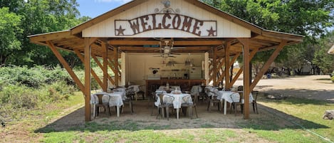 A wooden pavilion with a 'WELCOME' sign, tables with white tablecloths, and a kitchen area.