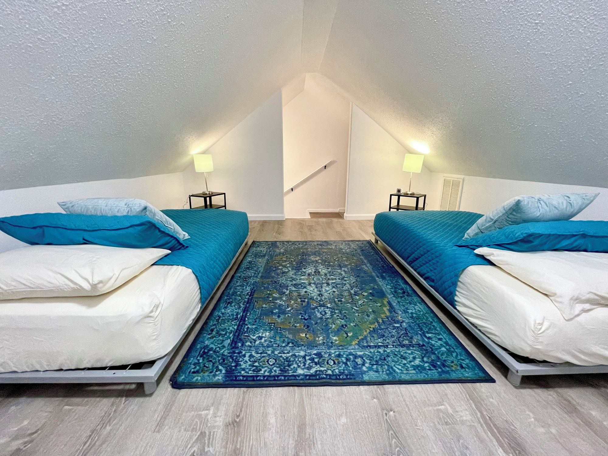 A room with two beds, a blue rug, and a staircase leading to an upper level.