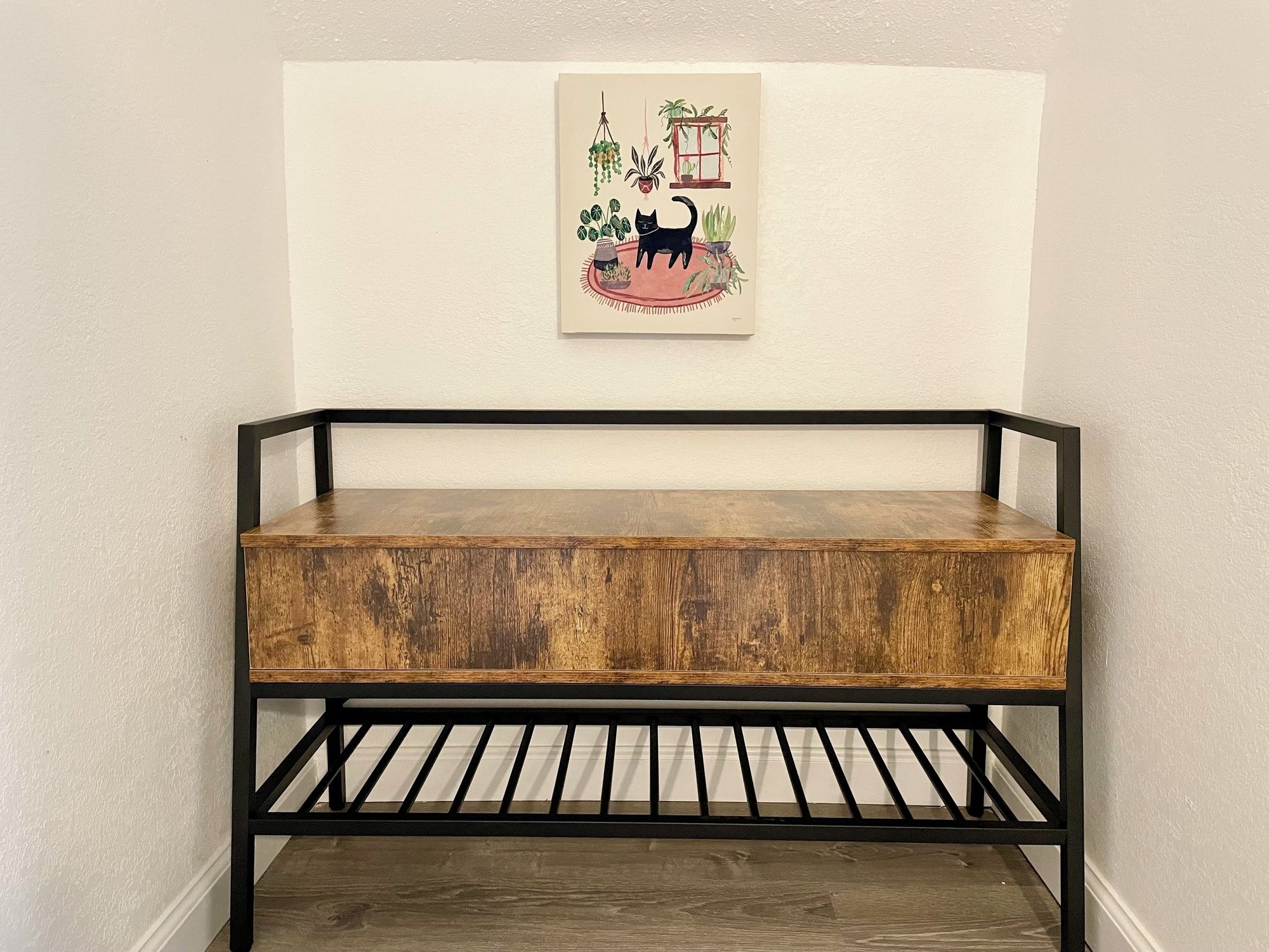 Un banc en bois doté d’une structure en métal noir, placé contre un mur sur lequel est accrochée une œuvre d’art encadrée.