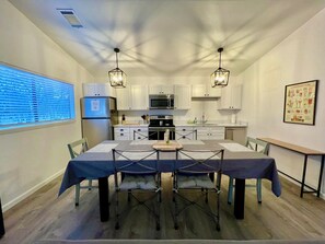 Cocina compacta con armarios blancos, microondas, estufa y refrigerador. Hay una mesa de comedor con sillas y una ventana con persianas.