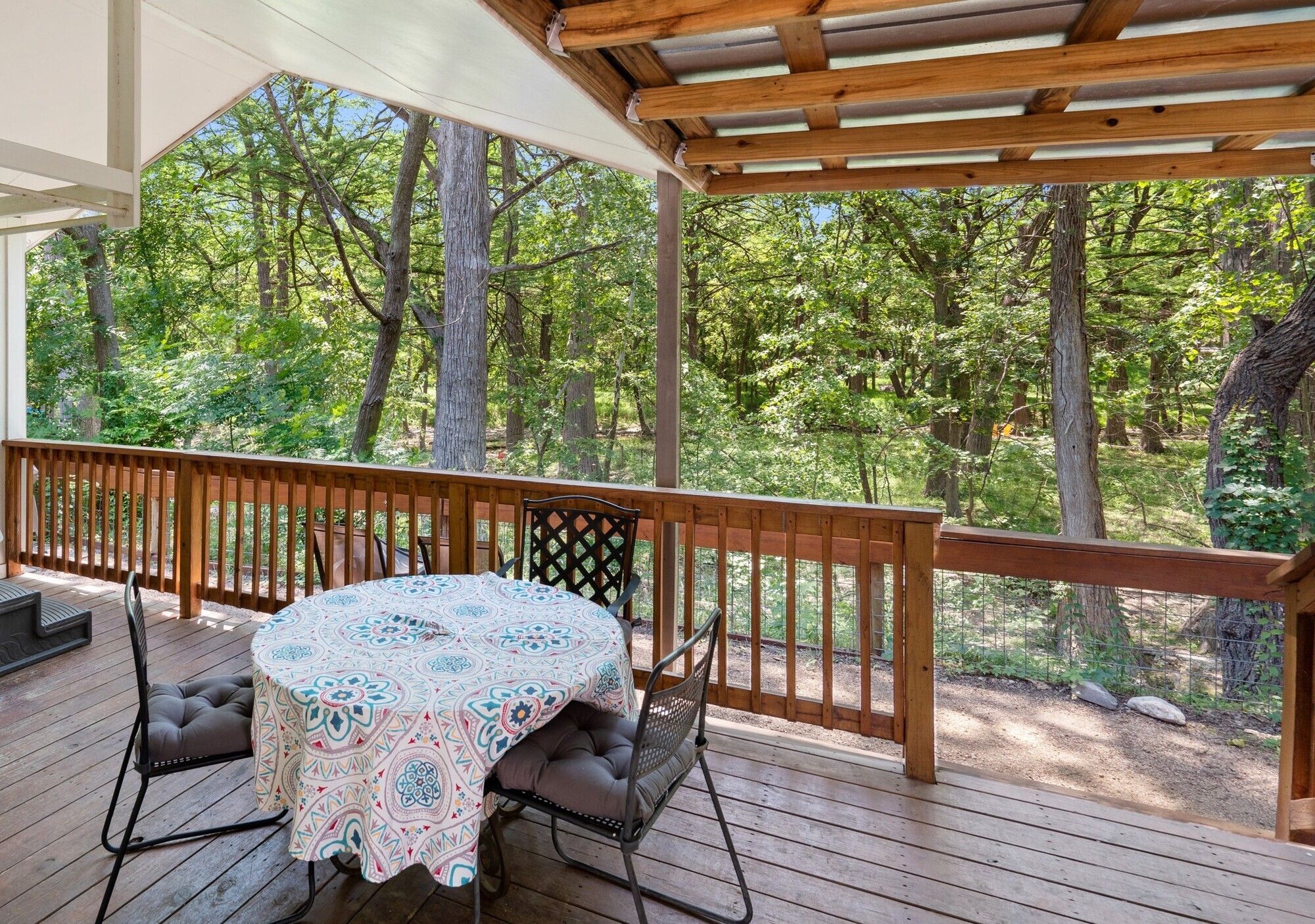 An outdoor deck with a table and chairs, surrounded by a wooden railing, overlooking a forested area.