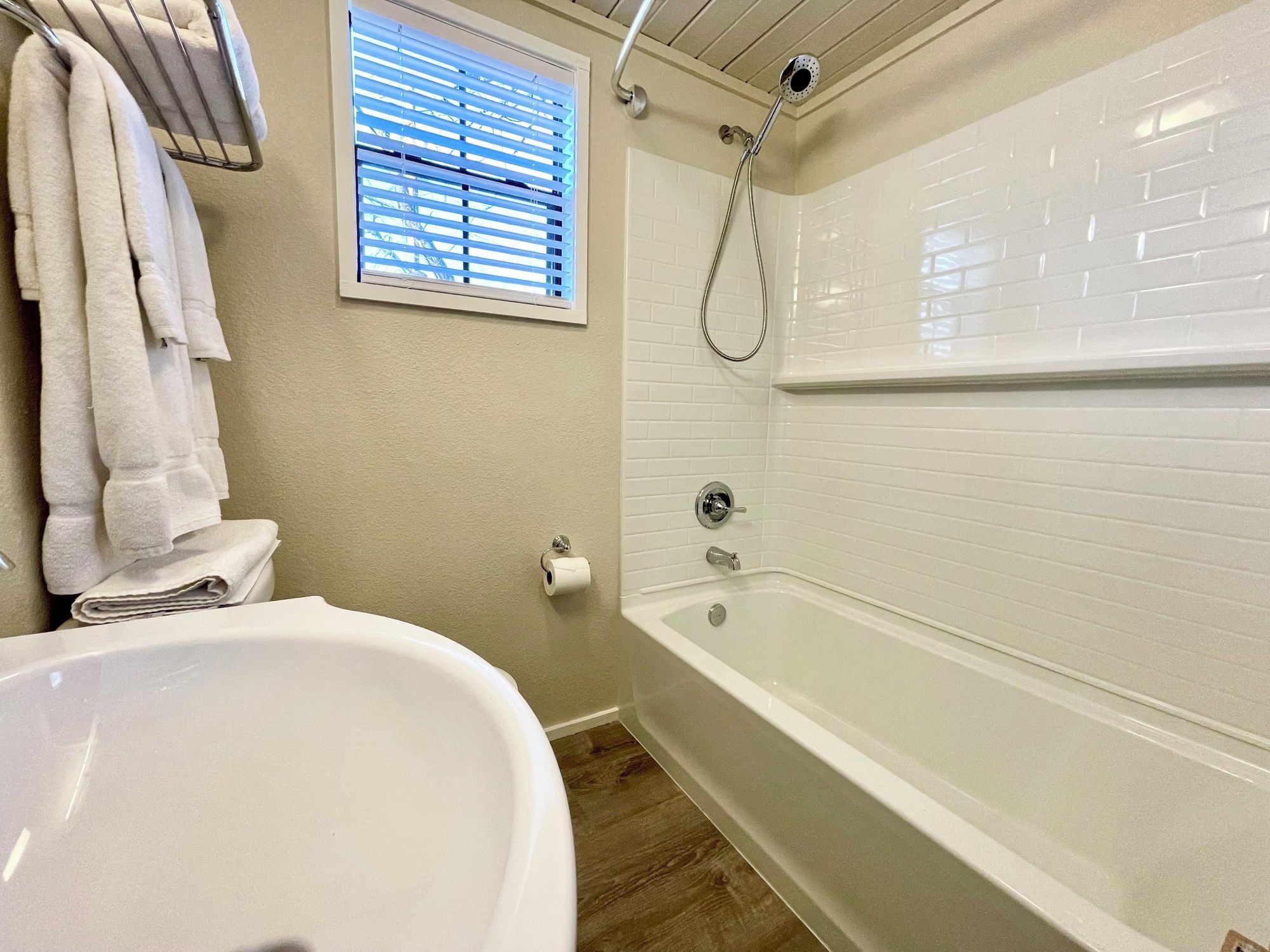 A bathroom with a white bathtub, a toilet, a towel rack, and a window with blinds.