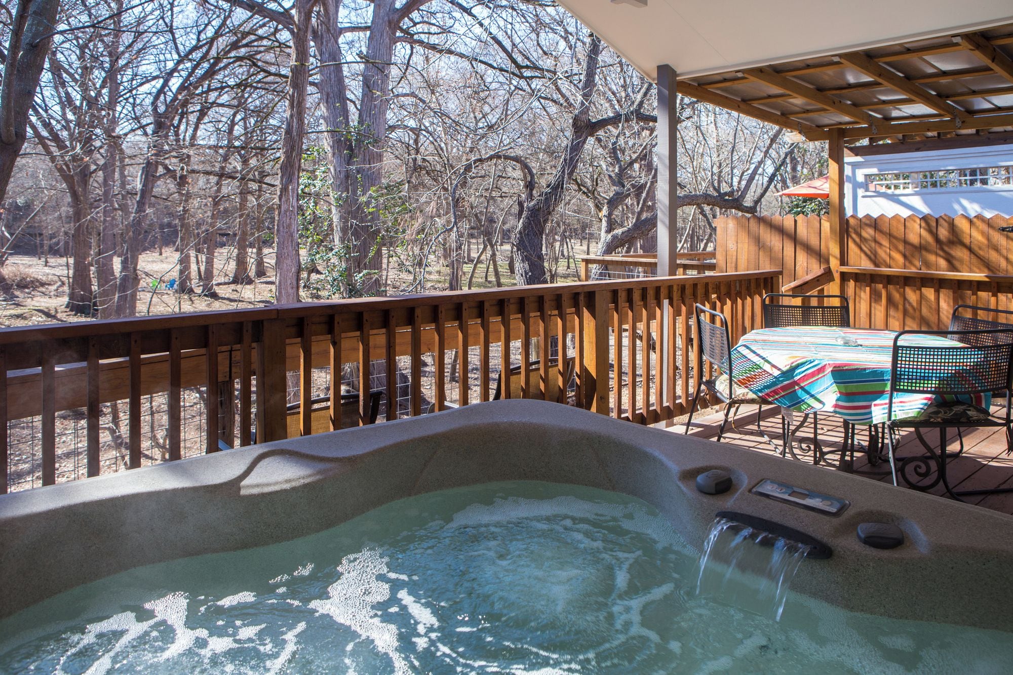 A hot tub with a wooden deck, a table with chairs, and a view of trees.