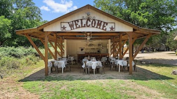 A wooden pavilion with a 'WELCOME' sign, tables with white tablecloths, and a kitchen area.