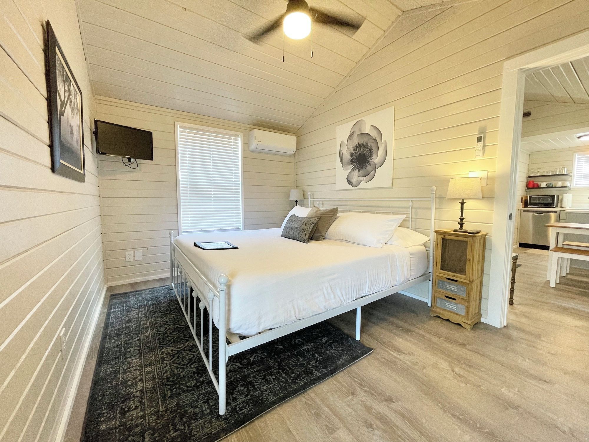 A bedroom with a bed, a bedside table, a lamp, a wall-mounted TV, and a window with blinds.