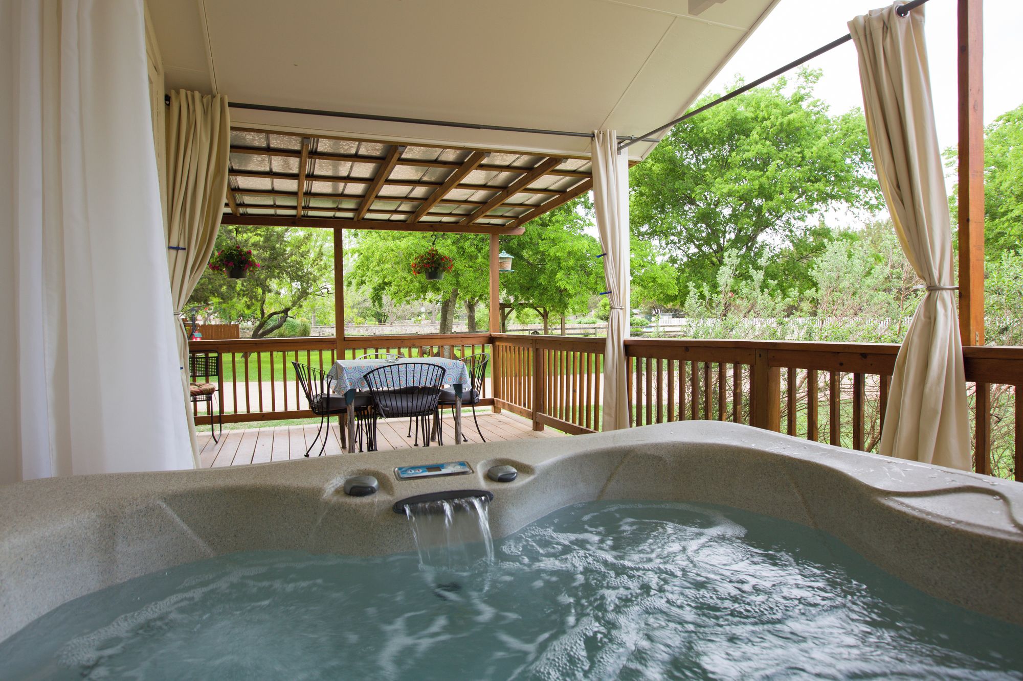 A hot tub with a wooden deck and a table with chairs, surrounded by a pergola with curtains.
