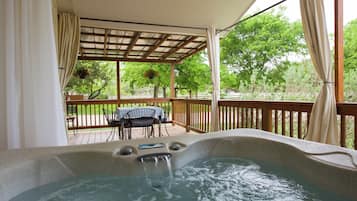 A hot tub with a wooden deck and a table with chairs, surrounded by a pergola with curtains.