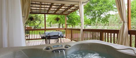 A hot tub with a wooden deck and a table with chairs, surrounded by a pergola with curtains.