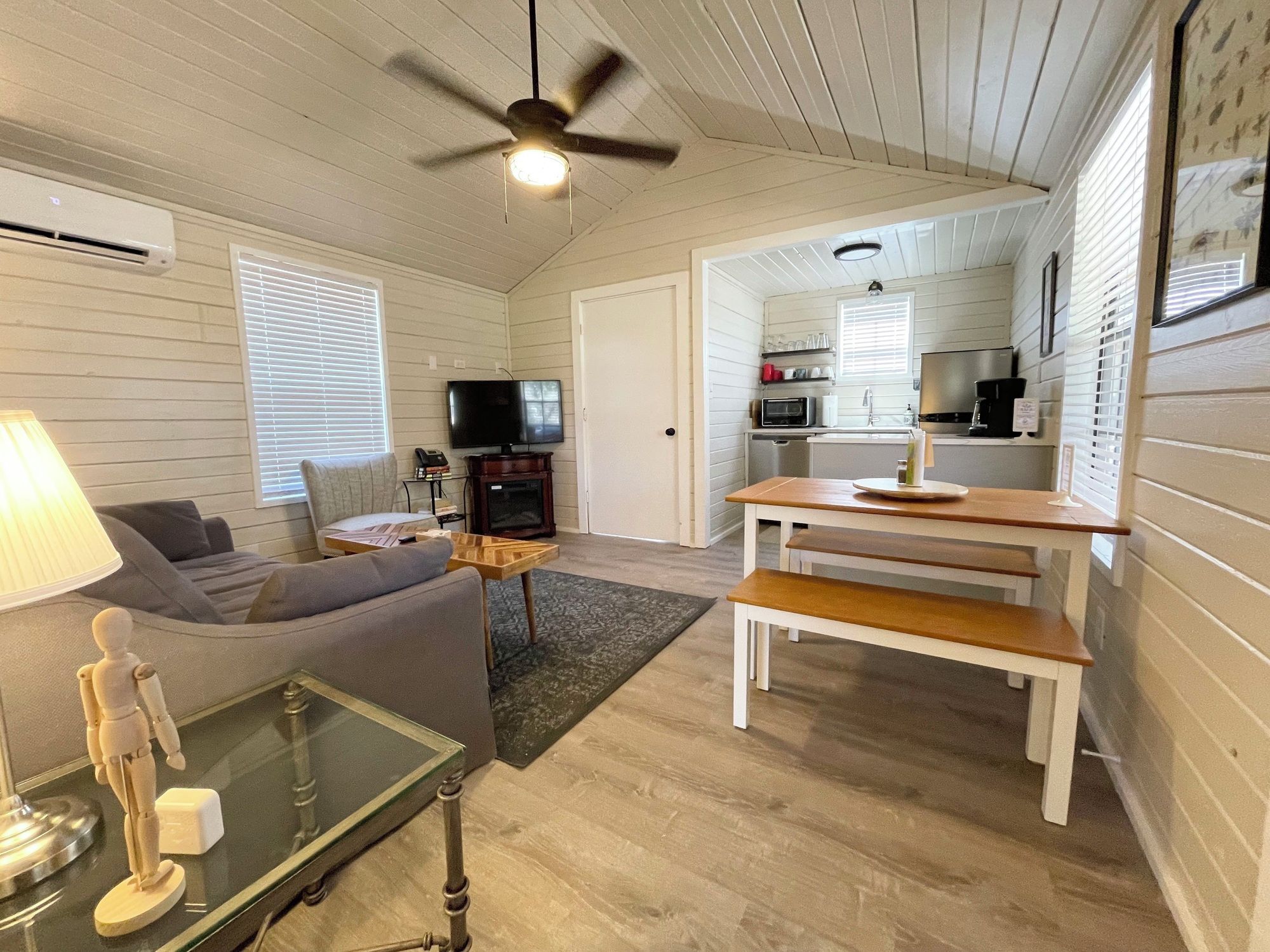 A compact living space with a sofa, a television, a dining table with benches, a ceiling fan, and a kitchen area with appliances.