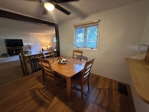 A dining area with a wooden table and chairs, a television on a brick wall, and a window with blinds. - GUNILLA's VILLA - The heart of downtown with privacy near LAKE NORMAN (Mooresville)