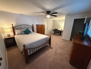 Ein Schlafzimmer mit einem Bett, einem Nachttisch, einer Kommode, einem Schreibtisch, einem Sessel und einem Deckenventilator.