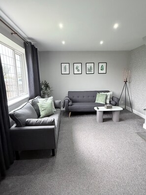 A modern living room with a grey sofa, a coffee table, and a floor lamp. There are four framed leaf prints on the wall. - House Sleeps 7 with Garden and Parking (Chester)