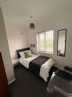A bedroom with a bed, bedside table, lamp, window with blinds, and a mirror on the wall. - House Sleeps 7 with Garden and Parking (Chester)