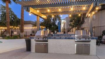 Un área de barbacoa al aire libre con dos parrillas a gas, un pérgola con luces colgantes y una piscina al fondo.