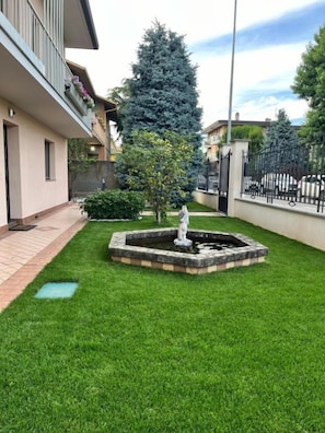 Un jardín bien cuidado con una fuente central, un sendero empedrado y un edificio con balcones.