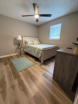 Ein Schlafzimmer mit einem Bett, einem Schreibtisch, einer Stehlampe, einem Deckenventilator und einem Fenster mit Jalousien.