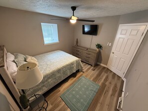 Une chambre à coucher comprenant un lit, une commode, un téléviseur, un ventilateur de plafond, une fenêtre et une porte.