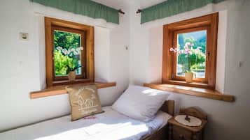 Pequeña habitación con cama individual, cabecera y piecera de madera, una mesita de noche y dos ventanas con marcos de madera y cortinas verdes.