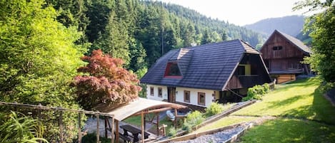 Una cabaña rústica con techo inclinado, rodeada por un camino de grava, y con un denso bosque y colinas al fondo.