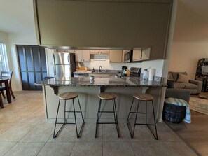 A modern kitchen with a central island, bar stools, stainless steel appliances, and a dining area with a table and chairs. - Coastal Retreat Near Parris Island in Beaufort (Beaufort)