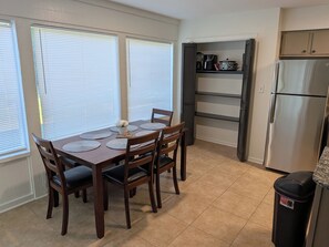 Uma sala de jantar com mesa posta para seis, com dois janelões com persianas, uma cozinha com frigorífico e estantes, e um cesto de lixo.