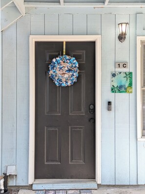 A dark brown door with a blue and white welcome wreath, a number plaque, and a wall-mounted light fixture. - Coastal Retreat Near Parris Island in Beaufort (Beaufort)