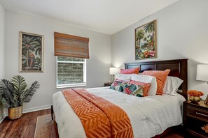 A bedroom with a bed, a window with blinds, a nightstand, a lamp, and two wall-mounted paintings. - Spacious, centrally located w/ Game room Tropical Tampa Florida Home! (Tampa)