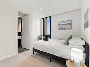 A bedroom with a bed, a nightstand, a lamp, a painting, and a view of the outdoors. - Melbourne CBD 2BR | Pool, Gym & City Views (Melbourne)