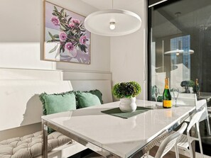 A modern dining area with a white table, green cushions, a small potted plant, and a bottle of champagne. - Melbourne CBD 2BR | Pool, Gym & City Views (Melbourne)