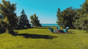 Ein grünes Areal im Freien mit vier blauen Adirondack-Stühlen, die um eine Feuerstelle aus Stein angeordnet sind.