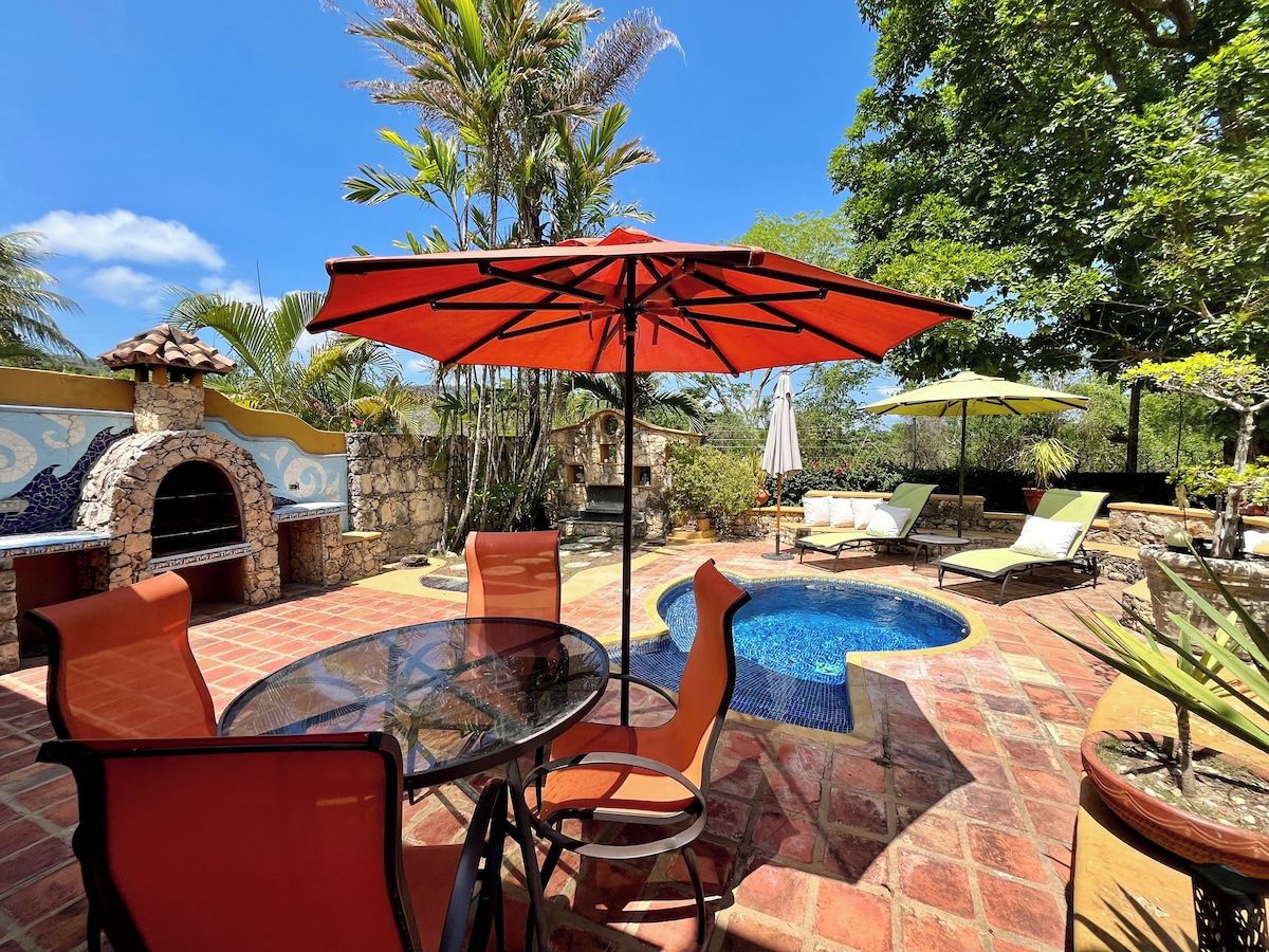 Un espace extérieur aménagé en terrasse, avec une table ronde en verre, des chaises orange, un parasol rouge, une cheminée en pierre et une piscine.