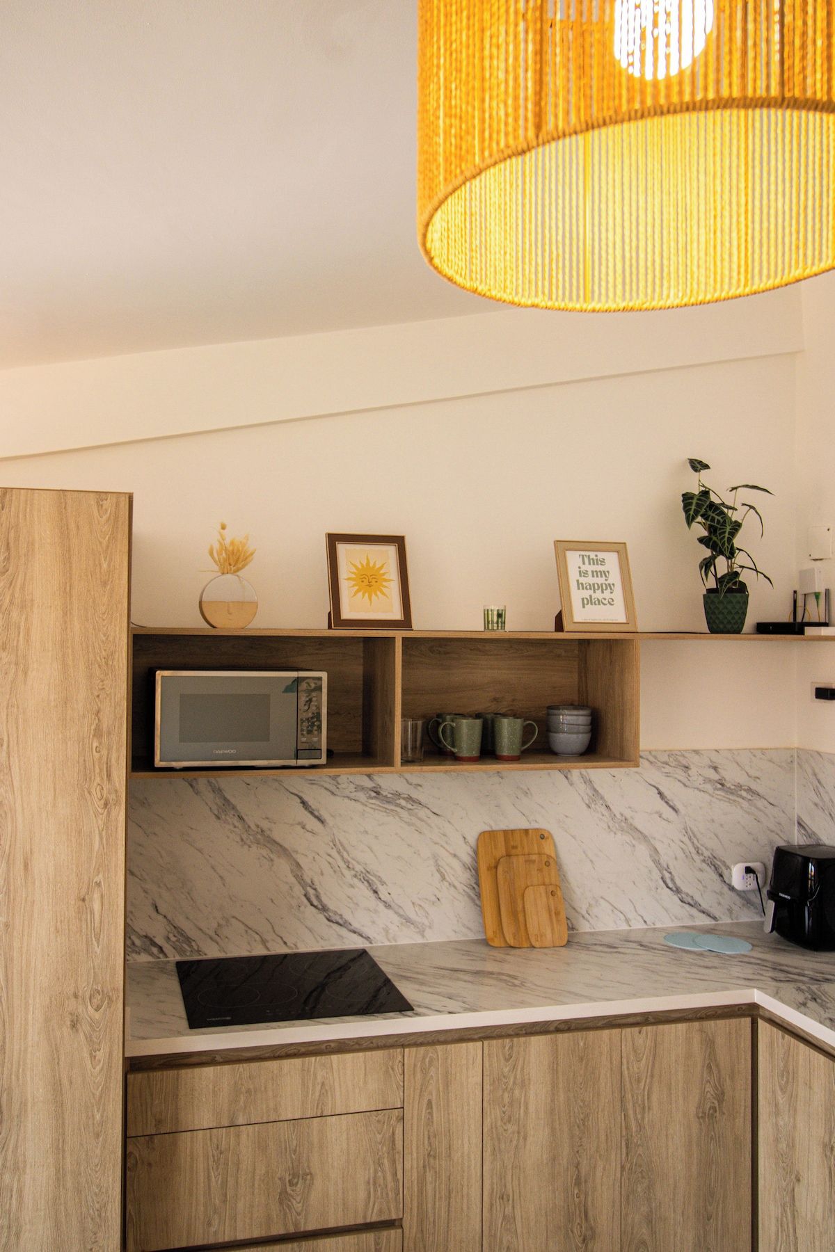 A modern kitchen with wooden cabinets, a marble countertop, a microwave, a cutting board, and a hanging light fixture.