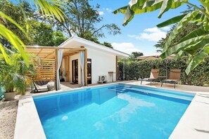 Ein Schwimmbecken mit Holzterrasse und Liegestühlen, umgeben von Grünflächen und einem weißen Gebäude mit einer überdachten Veranda.