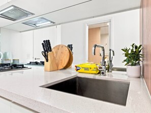 A modern kitchen with a stainless steel sink, a wooden cutting board, a set of knives, a yellow disinfectant wipes package, and a potted plant. - Sky-High South Yarra 1BR Apt w/ Pool, Gym & Views (South Yarra)