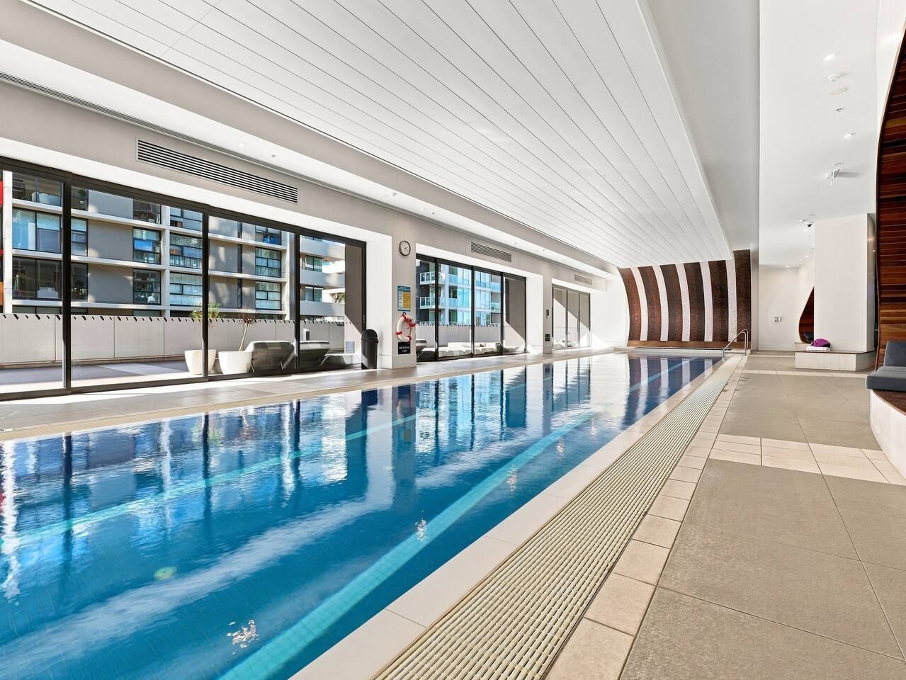 An indoor swimming pool with clear water, a tiled floor, and a modern architectural design with large windows and a high ceiling.