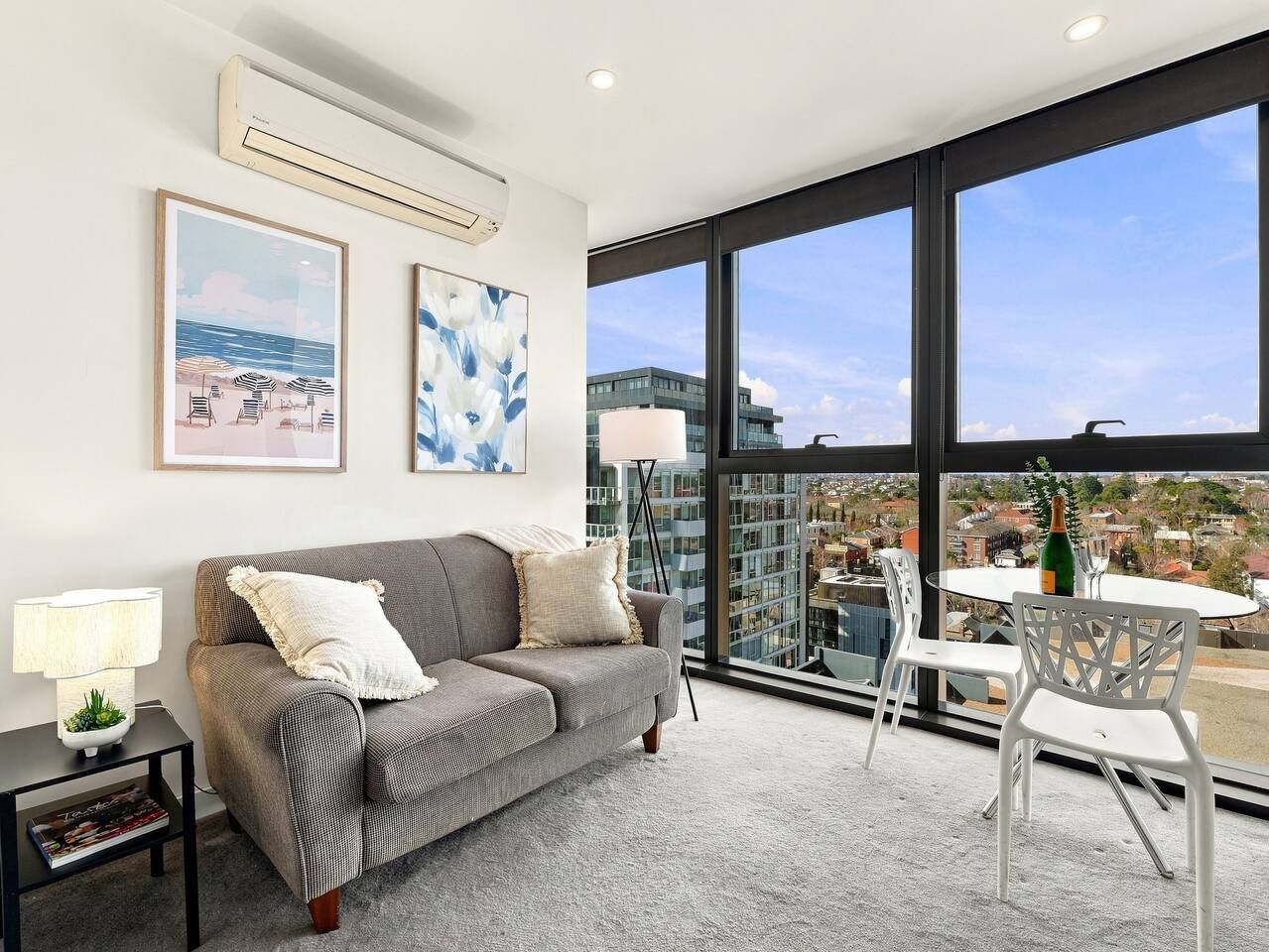 Una sala de estar moderna con un sofá gris, una mesita con una lámpara y un balcón con vistas a edificios.