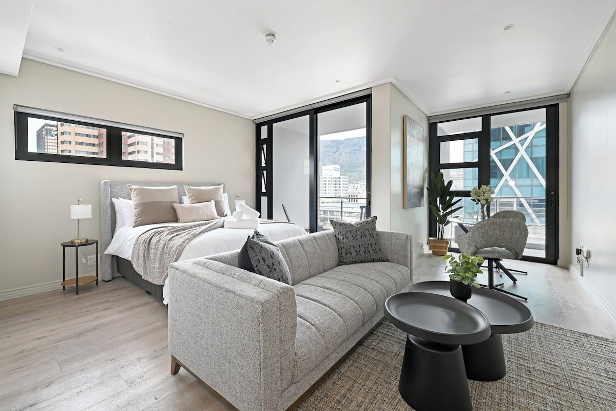 A modern bedroom with a large bed, a grey sofa, a small table, and a view of the city through large windows.