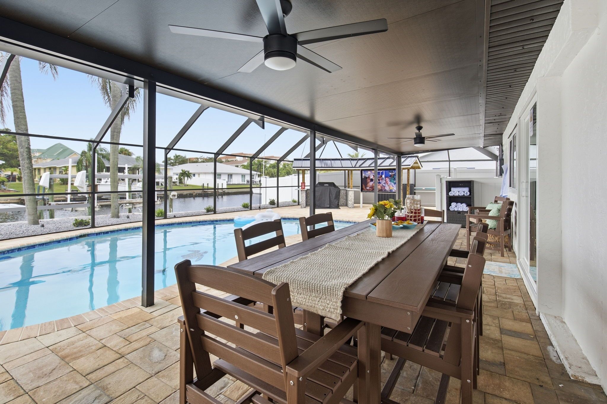 An outdoor pool area with a covered patio, wooden furniture, and a television screen.