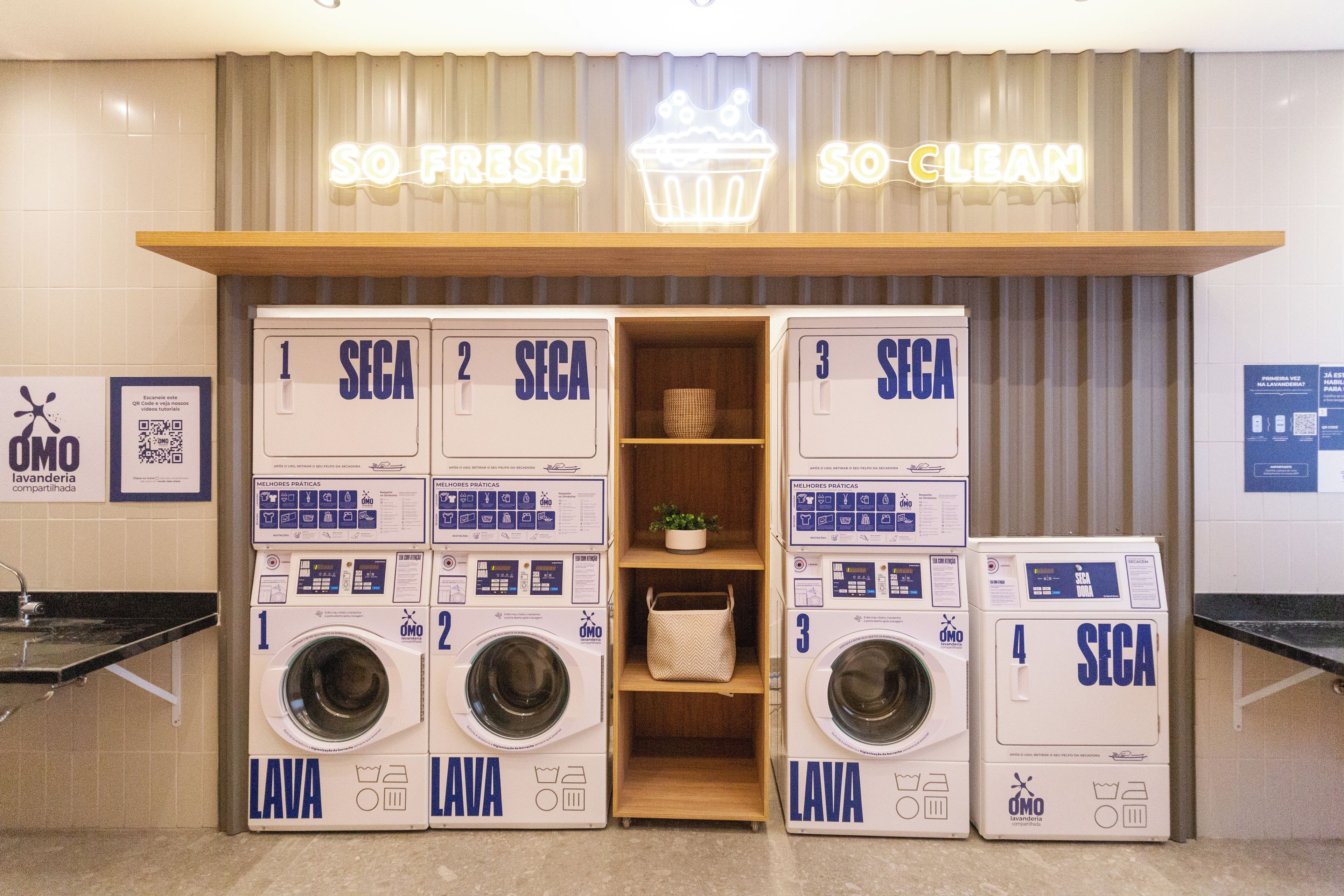 Un'area lavanderia con quattro lavatrici numerate da 1 a 4, uno scaffale con una pianta e un neon che riporta la scritta 'SO FRESH SO CLEAN'.