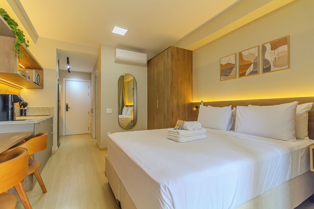 A hotel room with a bed, bedside table, and a desk with a chair. There is a mirror on the wall and a door leading to another room.