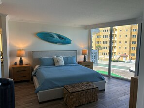 A bedroom with a bed, bedside tables, a nightstand, a decorative pillow, a wall-mounted surfboard, and a view of a building through a large window. - Toes-in-the-Sand - Oceanfront Views - New Smyrna Beach (New Smyrna Beach)