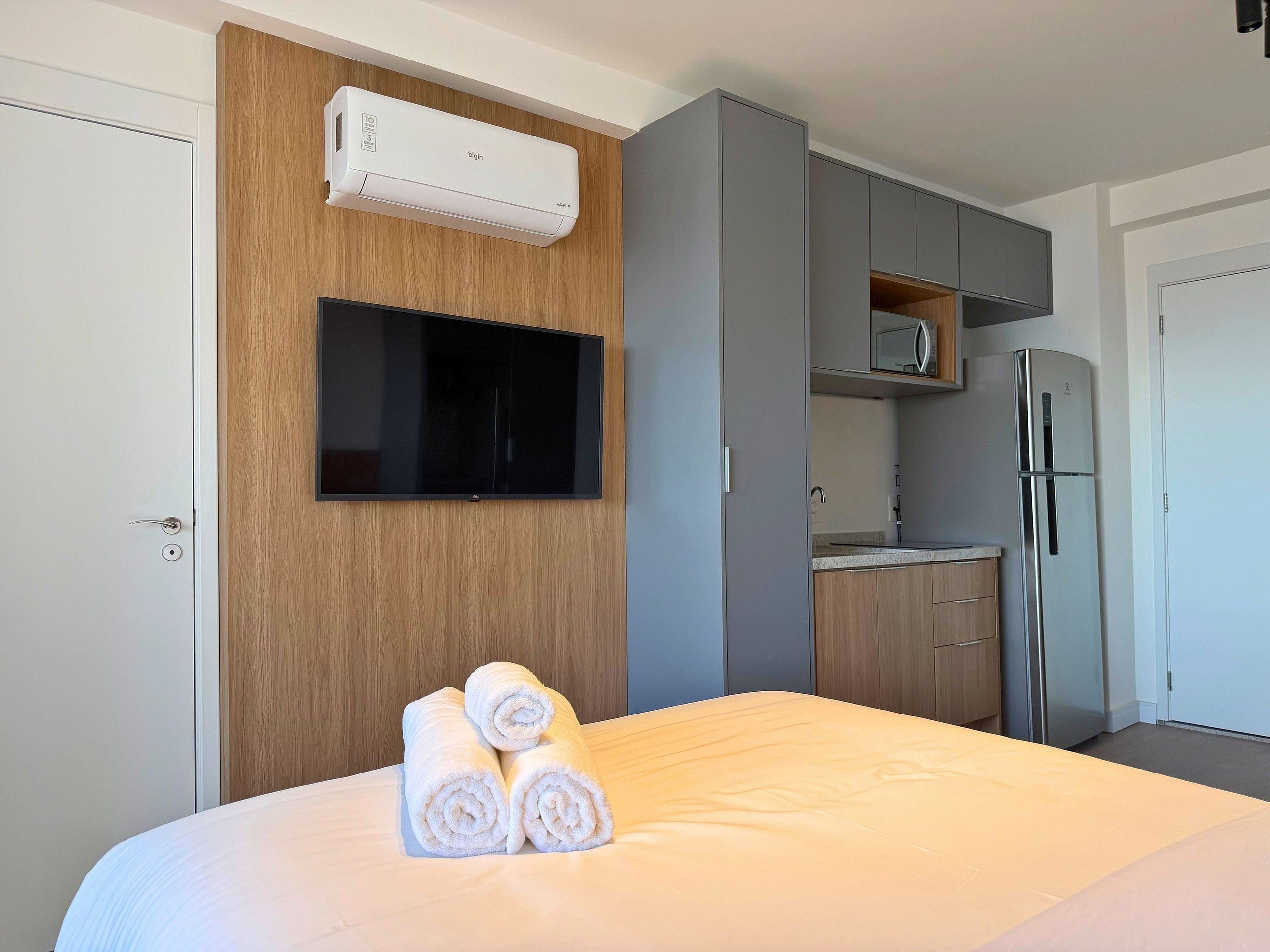 A hotel room with a bed, a flat-screen TV mounted on a wooden wall, a built-in air conditioner, a kitchen area with a microwave and refrigerator, and neatly rolled towels on the bed.