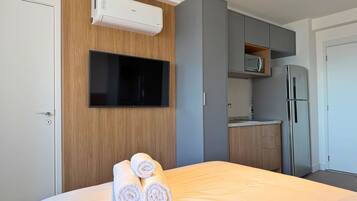 A hotel room with a bed, a flat-screen TV mounted on a wooden wall, a built-in air conditioner, a kitchen area with a microwave and refrigerator, and neatly rolled towels on the bed.