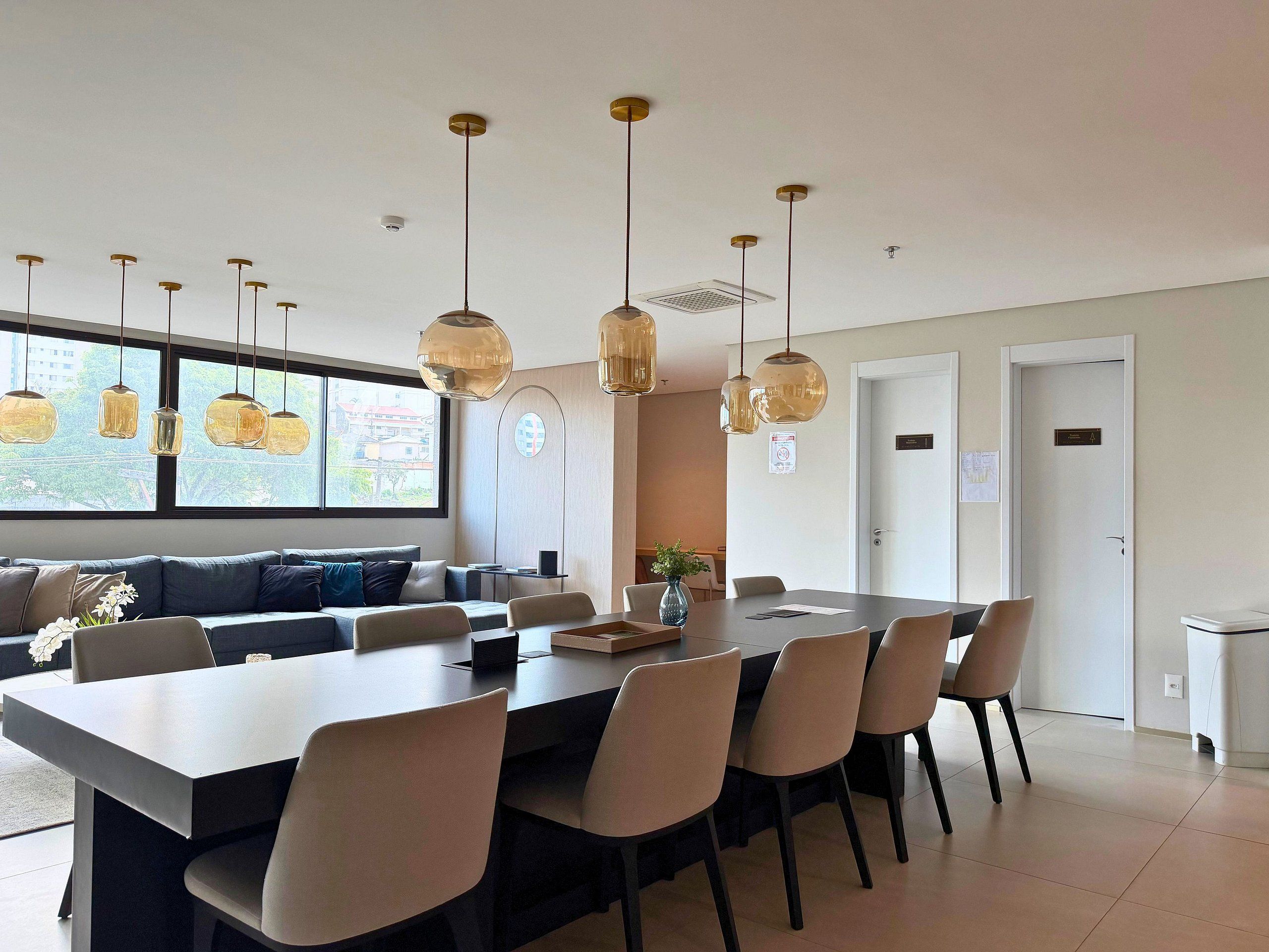 Uma sala de jantar moderna com uma mesa comprida e várias cadeiras, um sofá e iluminação pendente.