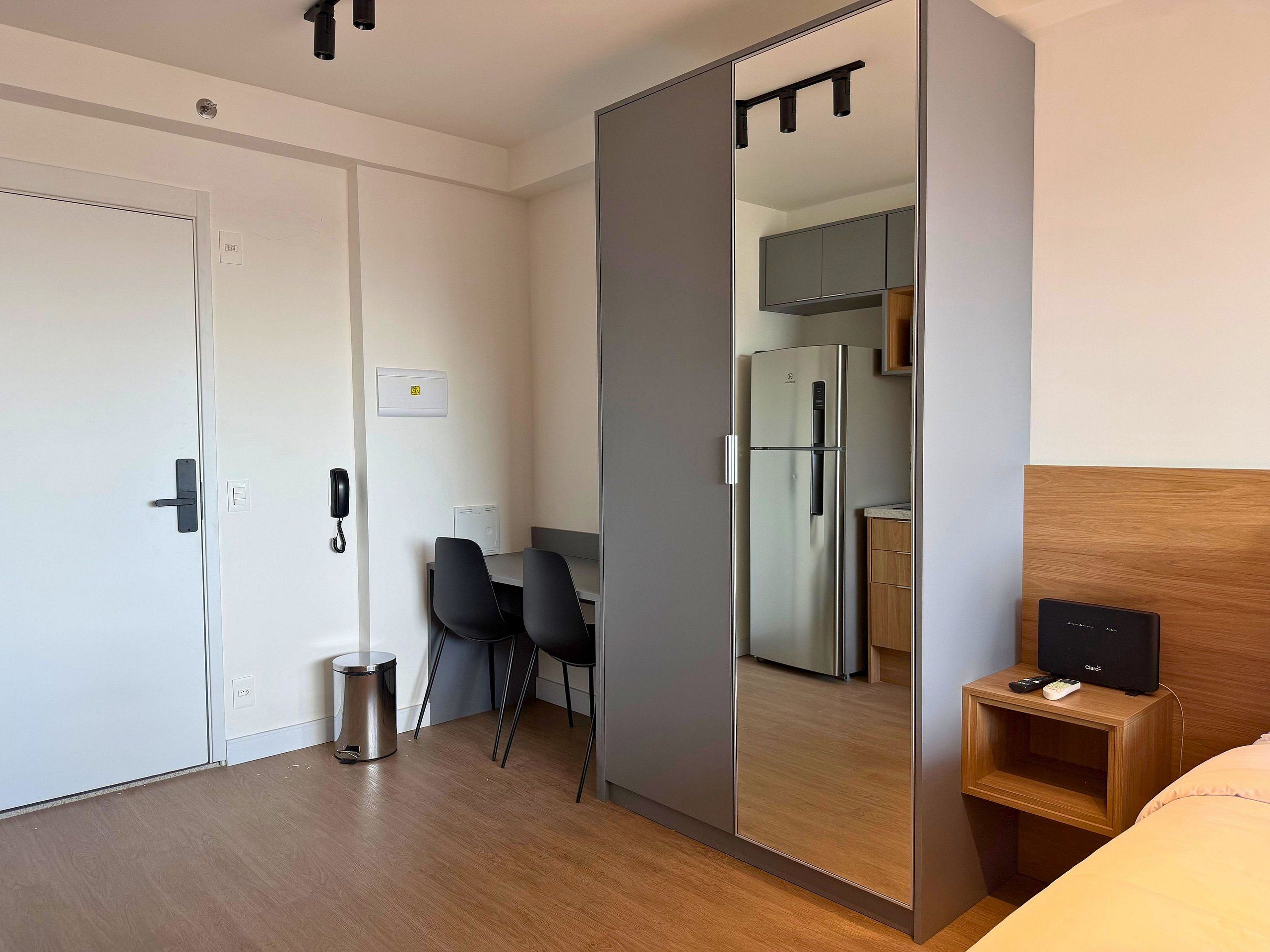 A hotel room with a sliding door closet, a small desk with a chair, a bed, a bedside table with a remote, and a kitchen area visible through the door.