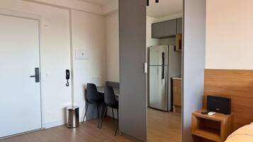 A hotel room with a sliding door closet, a small desk with a chair, a bed, a bedside table with a remote, and a kitchen area visible through the door.