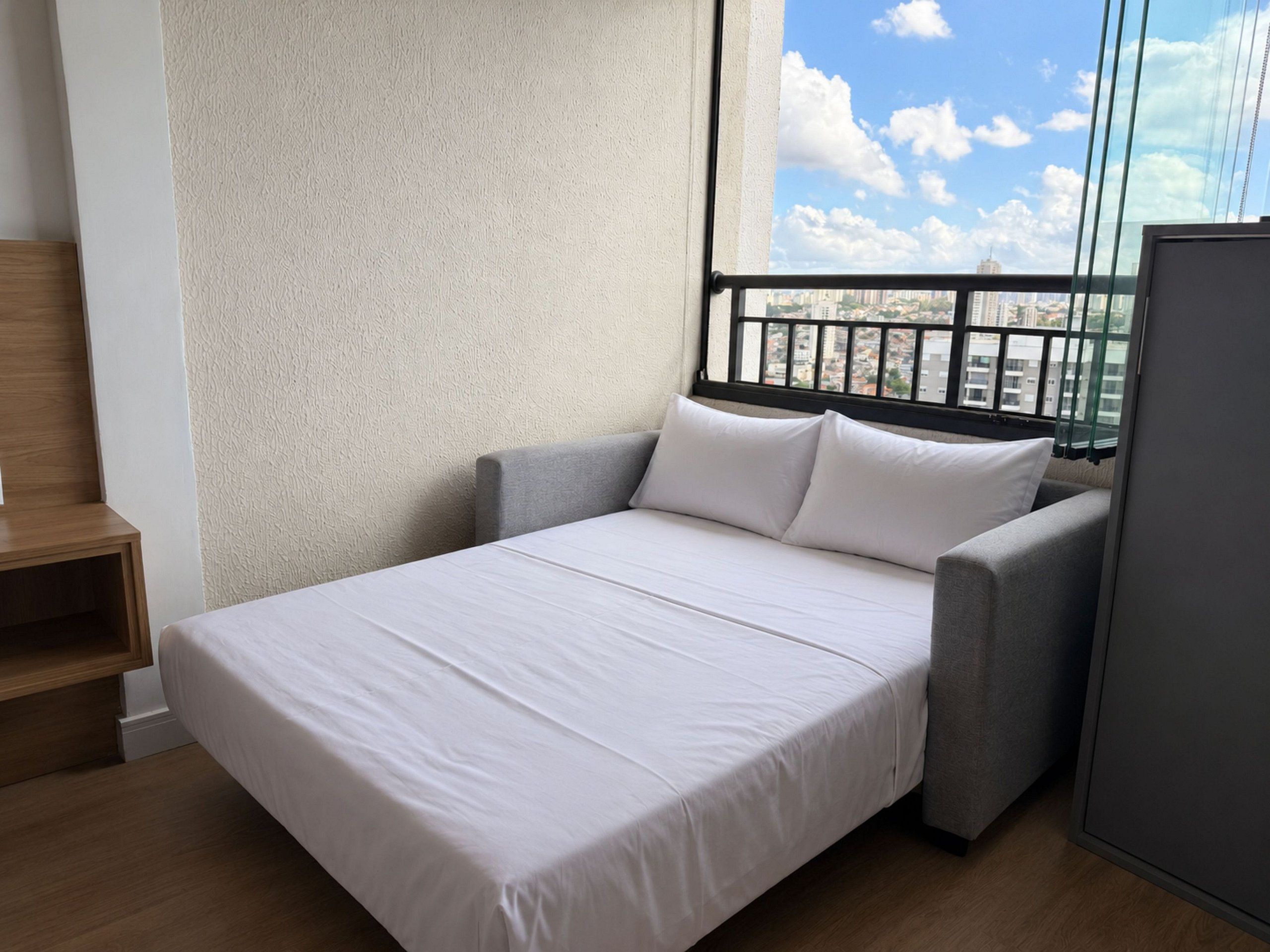 A single bed with a white mattress and gray headboard in a room with a view of a cityscape through a window with black railings.
