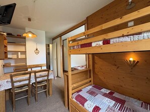 Een slaapkamer met een stapelbed, een houten bureau en stoelen, een hanglamp en een klein plankje.