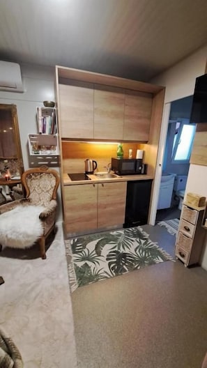A compact kitchen with wooden cabinets, a coffee maker, and a kettle. There is a patterned rug on the floor and a chair with a fluffy cushion.