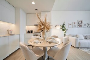 Une cuisine moderne avec des meubles blancs, un coin repas doté d’une table ronde pour quatre personnes et un salon avec un canapé et des plantes décoratives.